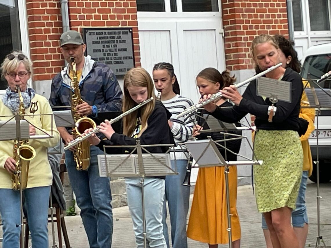"Des musiciens de l’Ensemble Instrumental Waremmien jouent dans la rue à Bleret, avec des flûtes traversières et saxophones devant un public curieux, lors du concert-apéro du 6 juillet 2025."