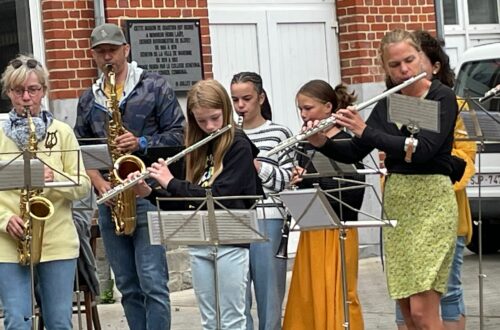 "Des musiciens de l’Ensemble Instrumental Waremmien jouent dans la rue à Bleret, avec des flûtes traversières et saxophones devant un public curieux, lors du concert-apéro du 6 juillet 2025."