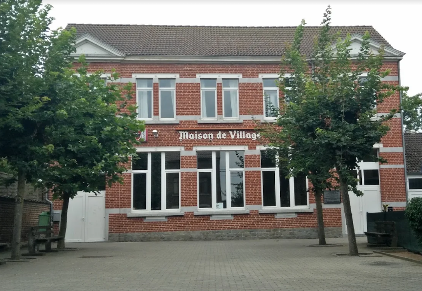 Photo de la Maison de Village de Bleret, un bâtiment en briques rouges avec des encadrements blancs, situé sur une placette pavée et bordée d’arbres.